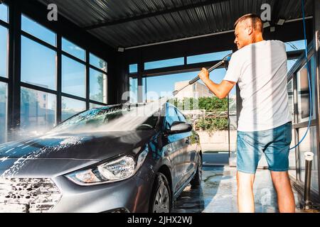 Lavage de voiture en libre-service. Un homme lave la mousse de sa voiture avec de l'eau d'un tuyau sous pression à un lavage de voiture en libre-service. Banque D'Images