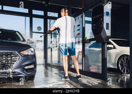 Lavage de voiture en libre-service. Un homme lave la mousse de sa voiture avec de l'eau d'un tuyau sous pression à un lavage de voiture en libre-service. Banque D'Images