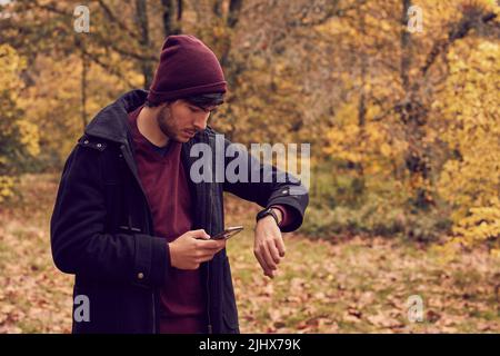 Blanc caucasien jeune homme synchronisation smartphone avec SmartWatch dans la forêt vêtu d'un manteau long et d'une casquette. Couleurs d'automne Banque D'Images