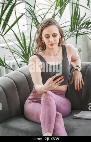 Femme sportive mince entraîneur de yoga posé sur un canapé dans des vêtements de sport avec téléphone mobile. Prendre des notes, planifier. Instructeur de fitness en vêtements de sport Banque D'Images