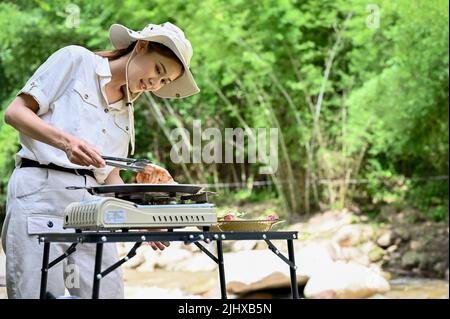 Charmante et décontractée, jeune femme asiatique préparant une partie de camping au barbecue, steak de porc grillé dans le magnifique terrain de camping. Banque D'Images