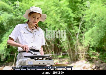 Une jeune femme asiatique heureuse et gaie aime préparer des plats, griller des steaks et des légumes sur son poêle à pique-nique portatif dans la forêt verte. Banque D'Images