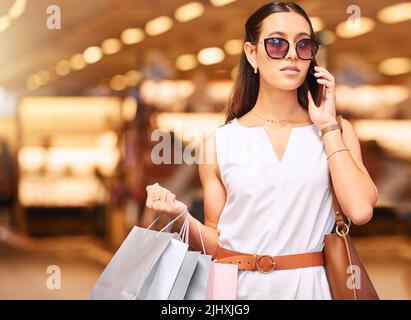 Une jeune femme élégante portant des lunettes de soleil et parlant sur un téléphone portable tout en portant des sacs pendant une frénésie de shopping. Une jeune femme brunette en appel avec elle Banque D'Images
