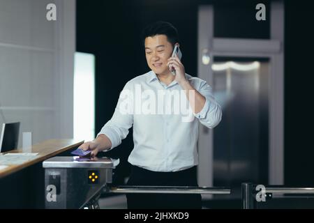 L'ouvrier entre dans le bureau, ouvre la porte avec une clé électronique avec une carte, un homme asiatique sourit et parle au téléphone Banque D'Images