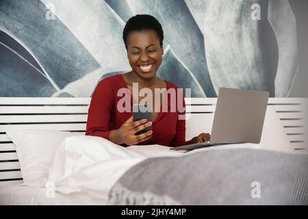 Femme africaine indépendante travaillant de sa chambre à la maison. Souriant regardant son téléphone avec ordinateur portable, allongé dans le lit Banque D'Images