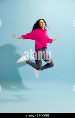 Jeune belle femme excitée dans une tenue de style décontracté saut isolé sur fond bleu. Concept de sport, beauté, art, mode, émotions humaines Banque D'Images