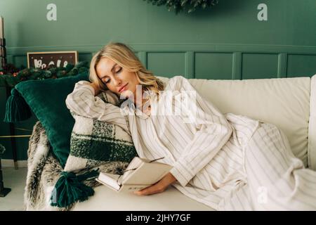 Une femme en pyjama est couché sur un canapé confortable dans un salon décoré pour Noël lire un livre. Banque D'Images