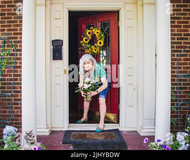 Jolie femme sénior en short et tee-shirt à la porte de la maison en brique avec des piliers et une couronne de tournesol ramasse la livraison de fleur laissée sur le porche pour la naissance Banque D'Images