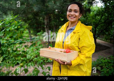 Portrait d'une femme africaine prospère et gaie inspirée jardinière dans un imperméable jaune souriant regardant l'appareil photo. Banque D'Images