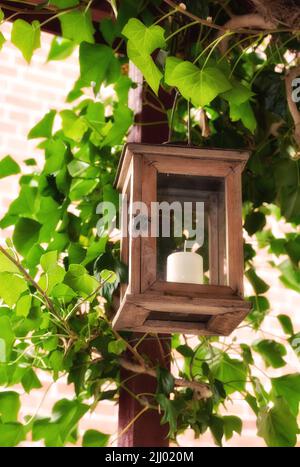 Bougie allumée dans une lanterne entourée de feuilles vertes dans un jardin ou un patio à l'extérieur. Cadre en bois suspendu sur le toit pour la décoration et la chaleur dans un Banque D'Images