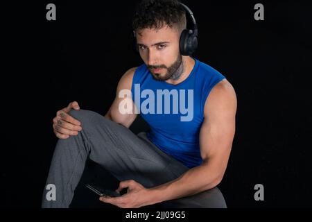 Beau homme avec tatouages dans son 20s dans la salle de gym avec téléphone et écouteurs, isolé sur fond noir Banque D'Images