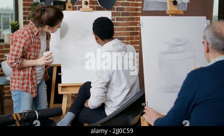 Jeune femme expliquant la leçon artistique à l'étudiant en cours d'art. Homme créatif utilisant un crayon et une toile avec chevalet, dessin vase chef-d'œuvre pour apprendre de nouvelles compétences en atelier avec enseignant. Banque D'Images