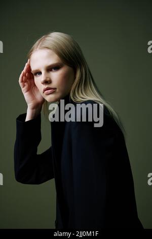 Un portrait rapproché d'une blonde regardant par-dessus son épaule dans une veste noire posant dans un studio sur un fond vert. Le concept de mode élégant Banque D'Images