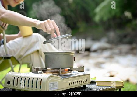 Gros plan, une campante femme prépare son déjeuner près de la rivière, de l'eau bouillante dans un pique-nique de camping sur un poêle portatif. Concept Camping Banque D'Images