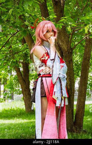 Costume d'anime dans le parc extérieur. Personnage de la déesse de jeu d'ordinateur avec des cheveux roses YaeMiko. Fille habillée comme héros d'anime se tient près arbre été Holida Banque D'Images