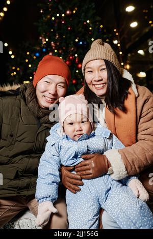 Photo verticale moyenne de parents asiatiques joyeux avec leur petite fille qui passe du temps à l'extérieur le soir d'hiver en souriant à l'appareil photo Banque D'Images