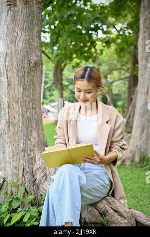 Une jeune femme asiatique attirante se détend dans le jardin verdoyant du parc, écrivant quelque chose sur son carnet ou lisant son roman préféré. Banque D'Images