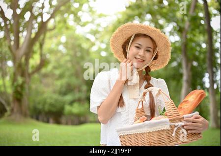 Jolie et attrayante jeune femme asiatique portant un chapeau de paille et tenant un panier de pâtisseries en osier dans le jardin vert. Concept pique-nique Banque D'Images