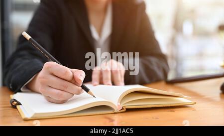 Une femme d'affaires en costume noir habillé écrivant quelque chose avec un crayon sur son carnet. Image rognée Banque D'Images