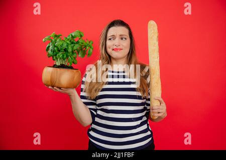 Confuse jeune femme à poil long dans une chemise rayée tenant un pot de fleur vert et une longue baguette française dans un studio rouge Banque D'Images