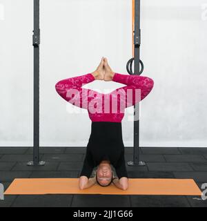 Jeune femme blonde exécutant la tête avec les genoux pliés et le pied ensemble pose yoga portant des leggings de yoga rose et une chemise noire dans une salle de gym. Banque D'Images