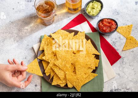 Tenir une main de femme de nachos, snack mexicain traditionnel servi avec de la guacamole, sauce chili rouge et la bière au ciment en arrière-plan. Vue d'en haut. Banque D'Images