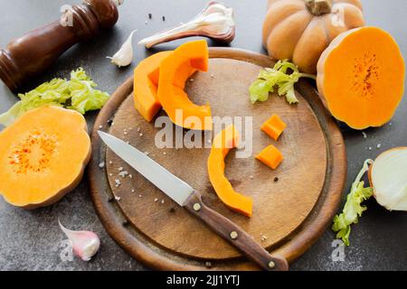 Potiron frais et brillant sur une planche à découper en bois avec couteau. Céleri et ail, oignons et grains de poivre épars et sel avec moulin à épices. Banque D'Images