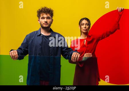homme curly et femme à la mode tenant différentes formes de verre tout en posant isolé sur jaune Banque D'Images