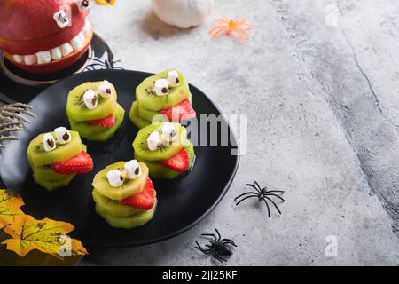 Monstres de kiwi vert effrayants pour Halloween. Gâteries d'Halloween à base de fruits sains. Halloween Party kiwi, fraise, pomme et monster de guimauve sur le eston gris Banque D'Images