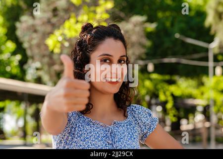 Jeune fille brunette souriante heureuse portant la robe d'été sur le parc de la ville, plein air positif, pouce vers le haut faisant l'excellent et signe d'approbation. Mise au point sélective Banque D'Images