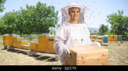 Jeune apiculteur femelle dans un uniforme tenant une boîte en bois sur l'apiculteur et les abeilles qui volent autour Banque D'Images