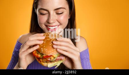 Une fille riante bante un gros hamburger savoureux. Femme souriante tenant un cheeseburger, mangeant, debout sur fond orange Banque D'Images