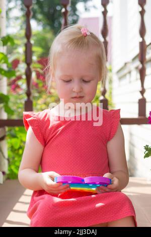 Mignon caucasien équitable cheveux fille dans la robe rose est de jouer et de montrer drôle tendance silicone antistress coloré jouet popit. Le jouet de violon Push Popper est le Banque D'Images