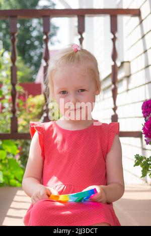 Mignon caucasien équitable cheveux fille dans la robe rose est de jouer et de montrer drôle tendance silicone antistress coloré jouet popit. Le jouet de violon Push Popper est le Banque D'Images