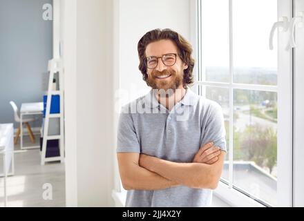 Portrait d'un homme d'affaires heureux dans un T-shirt et des lunettes debout près de la fenêtre du bureau Banque D'Images