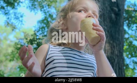 Une petite fille mignonne mange de la glace à l'extérieur. Portrait en gros plan d'une fille blonde assise sur le banc du parc et mangeant de la glace. Banque D'Images