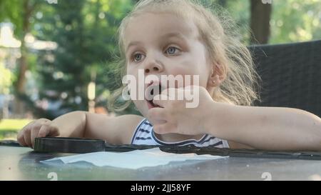 La petite fille a le goût du sel. Gros plan de la fille blonde prend le sel de la serviette avec son doigt et le goût tout en étant assis dans un café de rue sur le pair Banque D'Images