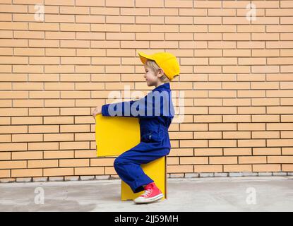 mignon garçon dans une combinaison bleue - l'uniforme joue avec des boîtes en carton jaune sur un fond de mur de brique. Livraison de colis, profession de postier, affaires Banque D'Images