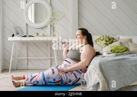 Femme parlant sur un téléphone portable par haut-parleur assis près du lit à la maison Banque D'Images