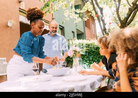 Femme souriante servant de la nourriture aux enfants debout près de l'homme à la table de salle à manger dans l'arrière-cour Banque D'Images