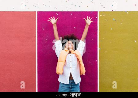 Fille avec les bras levés jeter confetti debout devant le mur coloré Banque D'Images