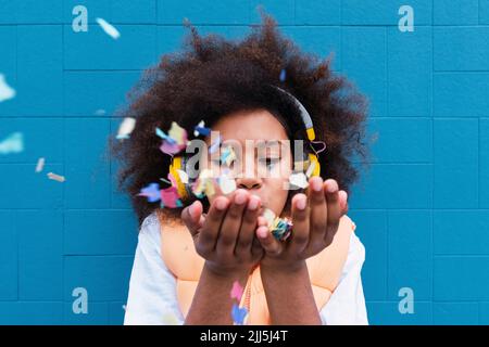 Fille portant un casque sans fil soufflant confetti devant le mur bleu Banque D'Images