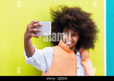 Bonne fille prenant selfie à travers le smartphone devant le mur jaune Banque D'Images