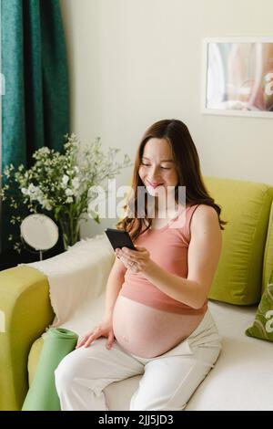 Bonne femme enceinte utilisant un smartphone sur un canapé à la maison Banque D'Images