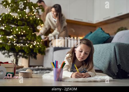 Fille écrivant la lettre au Père Noël avec les parents en arrière-plan à la maison Banque D'Images