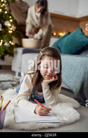 Fille avec la main sur le menton écrivant la lettre au Père Noël en étant couché à la maison Banque D'Images