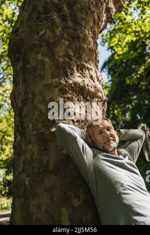 Homme âgé avec les mains derrière la tête, penché sur le tronc de l'arbre Banque D'Images