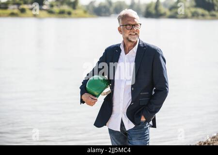 Homme senior souriant tenant un casque devant la rivière Banque D'Images