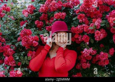 Femme souriante portant un chapeau rouge debout avec les yeux fermés au milieu des roses Banque D'Images