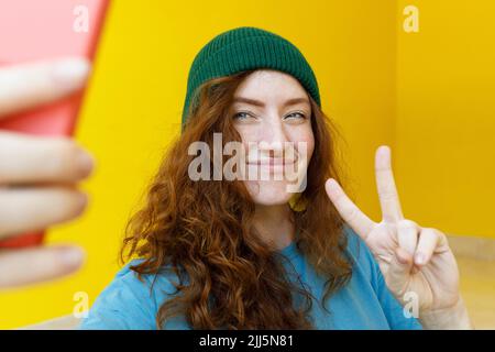 Femme souriante verte vêtue d'un chapeau en tricot emportant un selfie à travers un smartphone à la maison Banque D'Images
